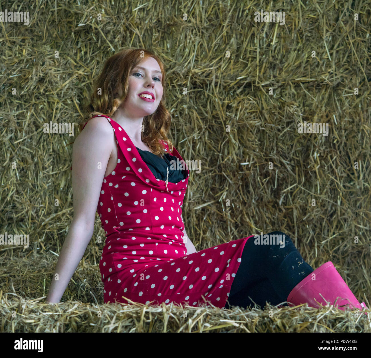 Testa rossa ragazza con red vintage polka dot abito rosa e gli stivali da pioggia si siede su balle di fieno in un accogliente fienile. Foto Stock