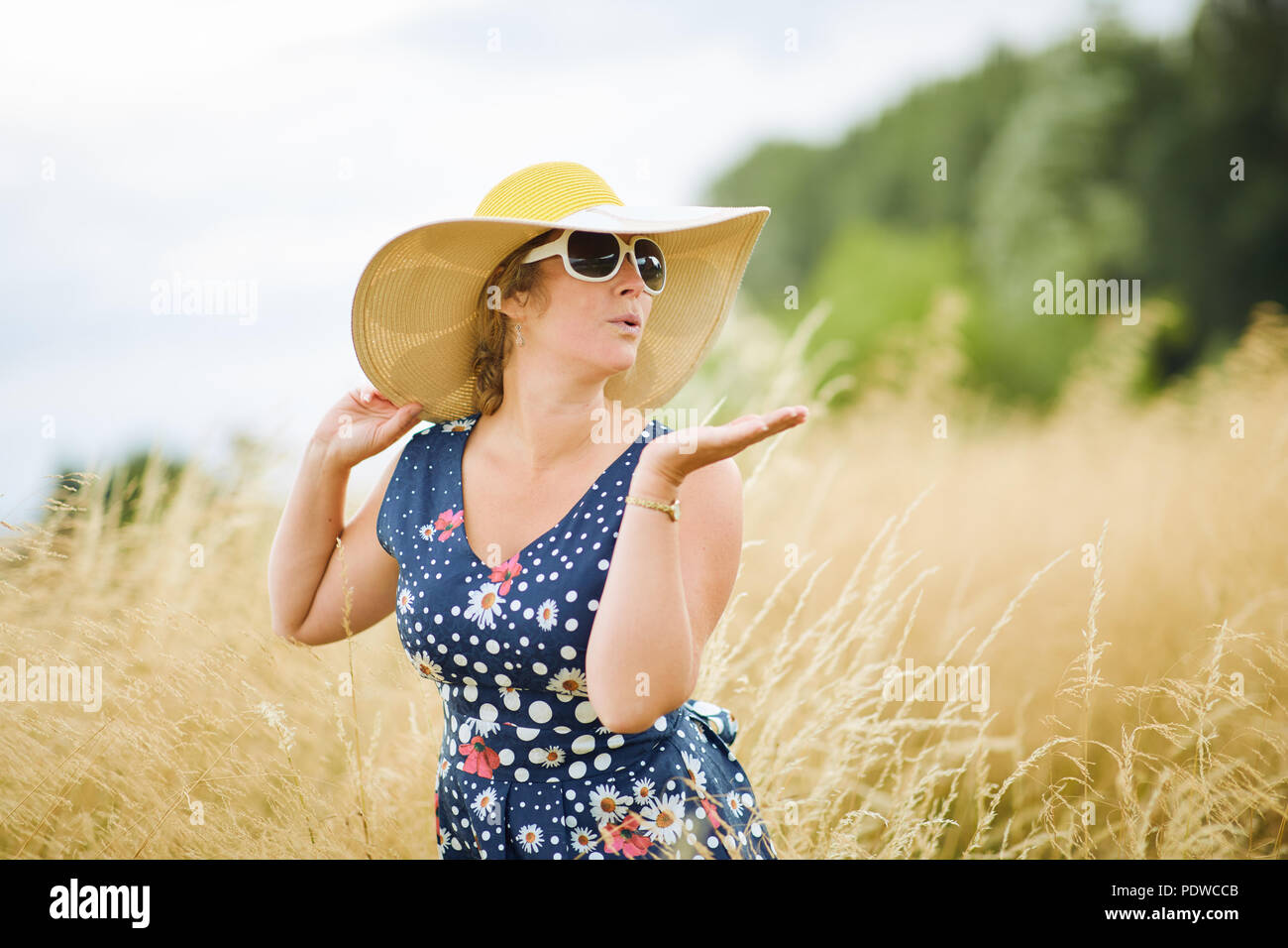 Donna di mezza età che indossa gli occhiali da sole bianco con una vasta colmato in piedi hat in lungo whispy golden erba mentre soffia un bacio su un luminoso giorno Foto Stock