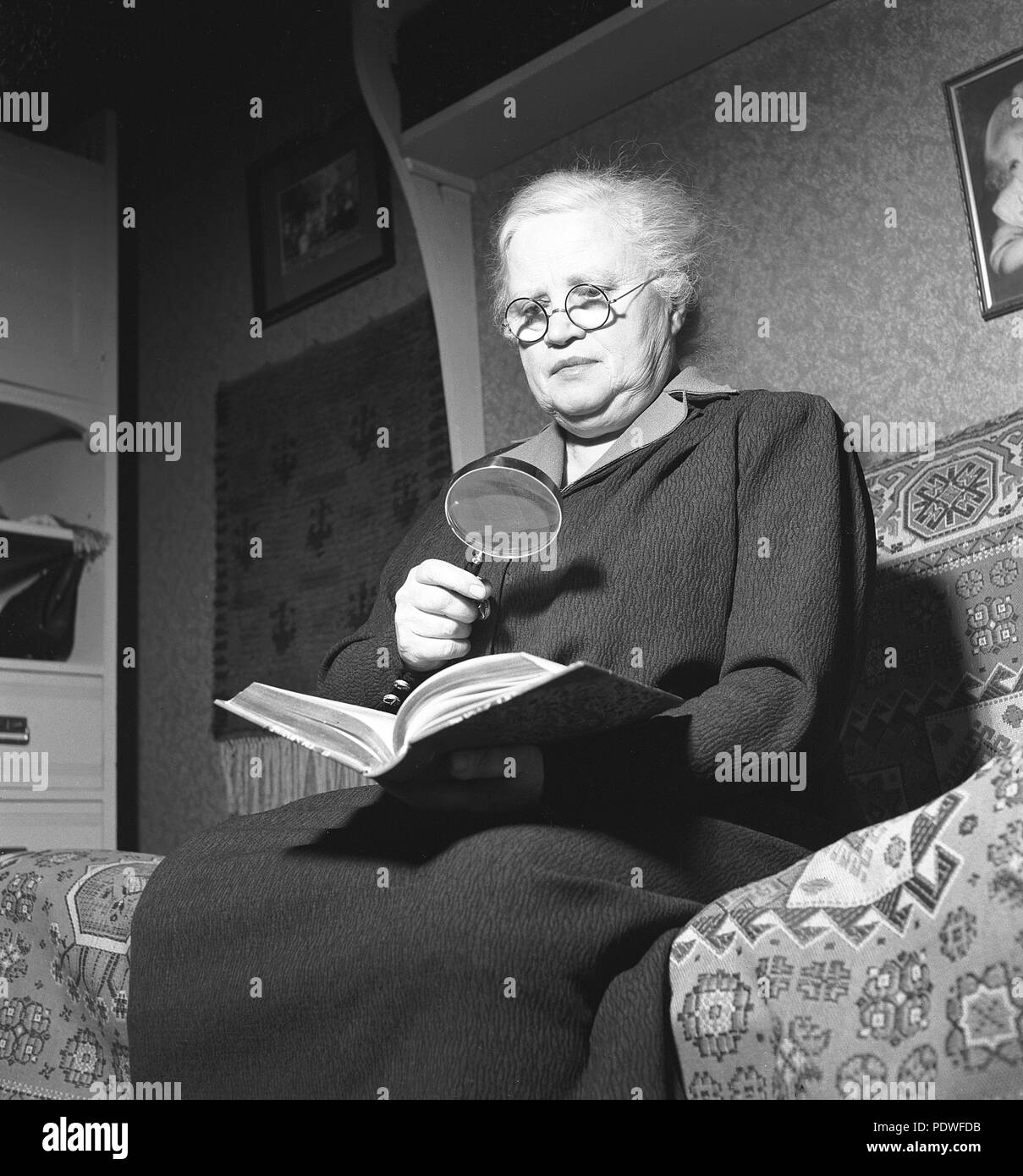 1940s donna con gli occhiali. Una vecchia Signora sta cercando in un libro usando entrambi i bicchieri e una lente di ingrandimento. La maggior parte delle persone associa la lente di ingrandimento con il carattere detective privato Sherlock Holmes creato da Conan Doyle. È anche un simbolo comune e icona per chiarire le funzioni di ricerca e di zoom in avanti i programmi di computer e su internet. 1942. Foto Kristoffersson/A84-6 Foto Stock