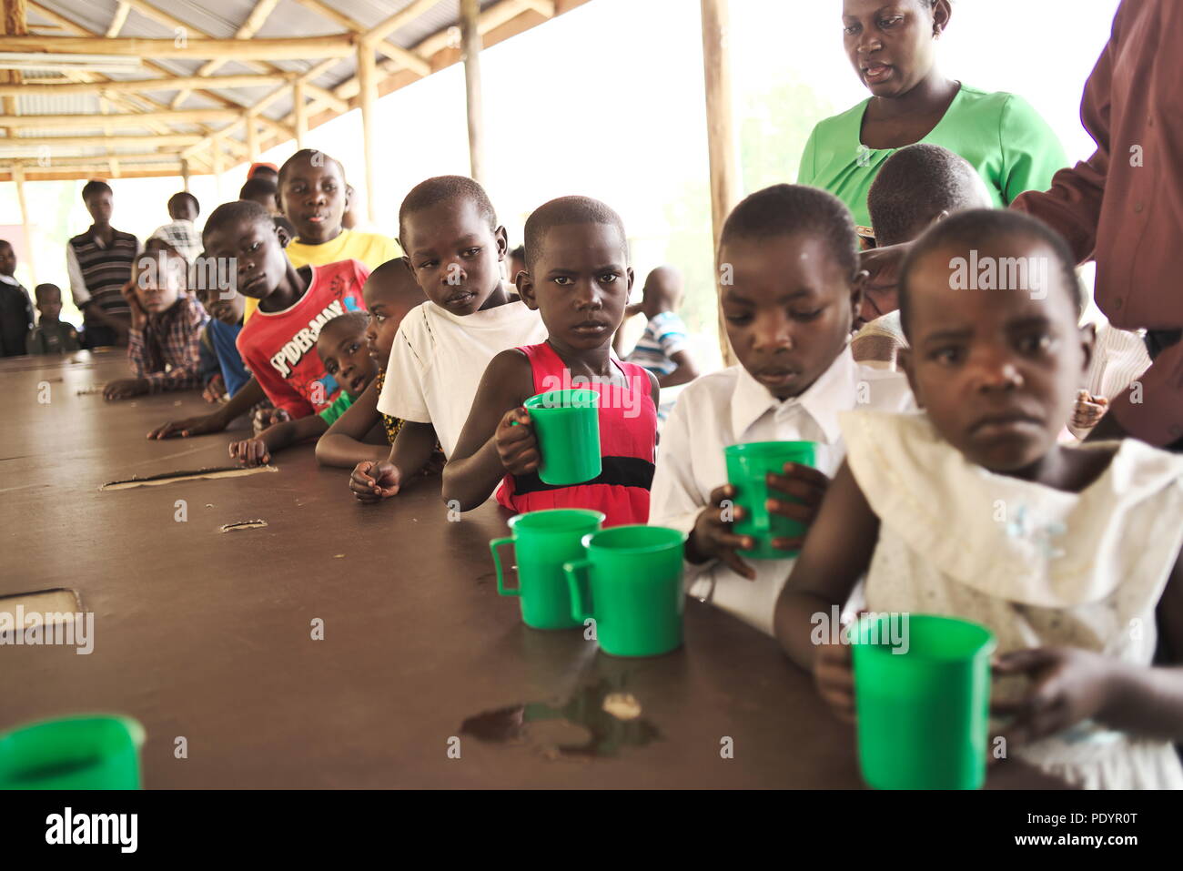 I bambini africani attendono in linea fuori da un ospedale ugandese in attesa di cibo e bevande con tazze di plastica e ciotole che guardano miserabili Foto Stock
