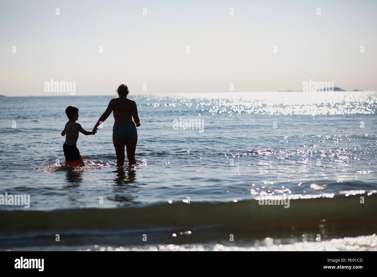 Madre e figlio divertirsi saltando le onde del mare nelle prime ore della sera mentre il sole tramonta ed è luccicante sulla pura acqua blu Foto Stock