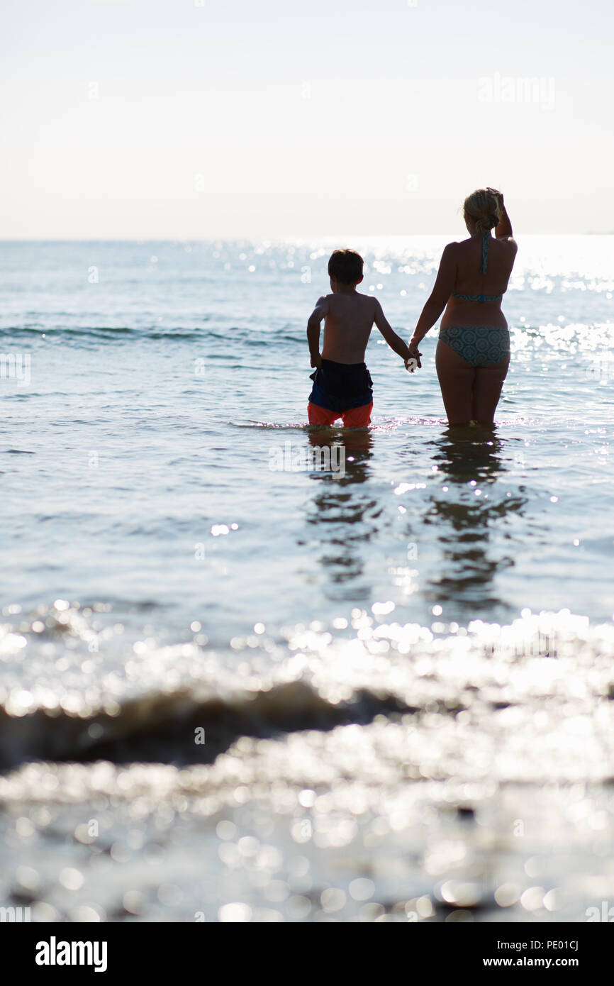 Madre e figlio divertirsi saltando le onde del mare nelle prime ore della sera mentre il sole tramonta ed è luccicante sulla pura acqua blu Foto Stock
