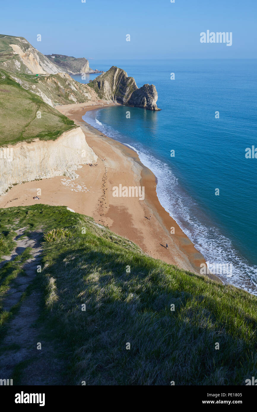 Guardando verso il basso dal verde clifftops erbose a Durdle porta dal South West Coast path, con chiare acque blu, una spiaggia di sabbia ed un cielo blu chiaro Foto Stock