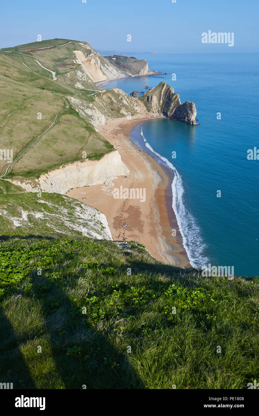 Guardando verso il basso dal verde clifftops erbose a Durdle porta dal South West Coast path, con chiare acque blu, una spiaggia di sabbia ed un cielo blu chiaro Foto Stock