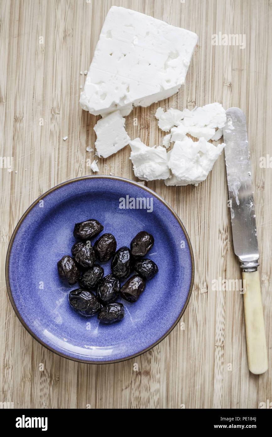 Il formaggio Feta, una ciotola di olive nere e un coltello su un pannello di legno Foto Stock