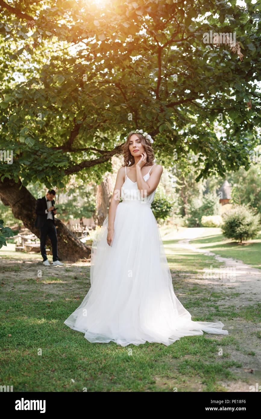 Giovane ragazza in un abito bianco nel prato. La donna in un bellissimo abito lungo in posa in giardino. Sposa mozzafiato in un abito da sposa. Foto Stock