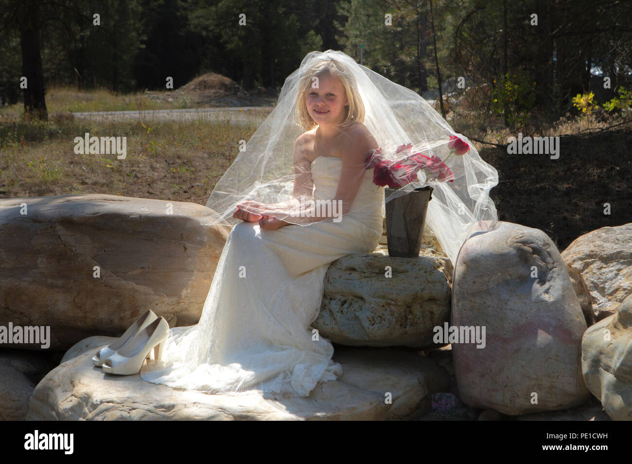 Fantasia, 8-9 anno attractie bionda, indossando zia di abito da sposa & velo. Seduti all'aperto sulla grande roccia accanto al vaso di rose rosse. Foto Stock