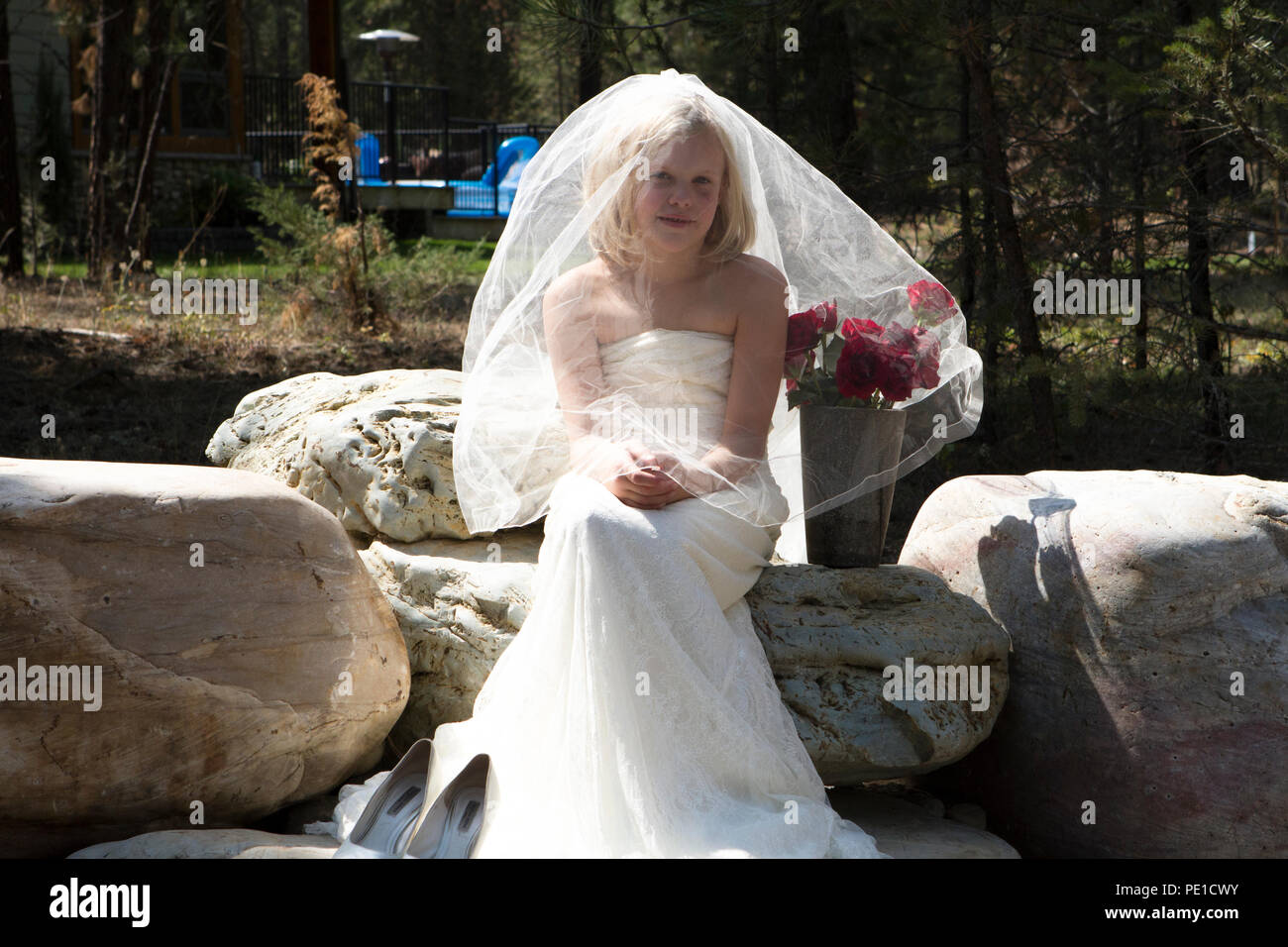 Fantasia, 8-9 anno attractie bionda, indossando zia di abito da sposa & velo. Seduti all'aperto sulla grande roccia accanto al vaso di rose rosse. Foto Stock