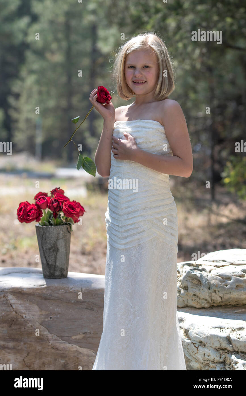 Fantasia, 8-9 anno attractie bionda, indossando zia di abito da sposa . In piedi all'aperto, tenendo una rosa rossa, accanto al vaso di rose rosse. Sorridente in telecamera Foto Stock