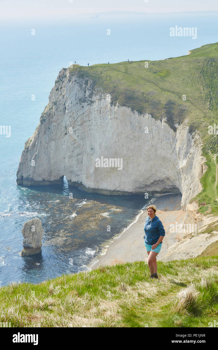 Donna in piedi su una banca erbosa in ore diurne con uno zaino, sulla costa sud dell'Inghilterra, guardando la telecamera con le bianche scogliere sul mare a t Foto Stock