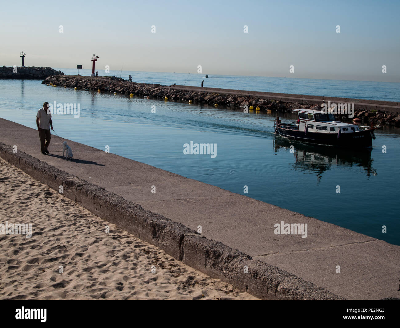 Un uomo cammina il suo cane e orologi una piccola barca che entra nel porto Foto Stock