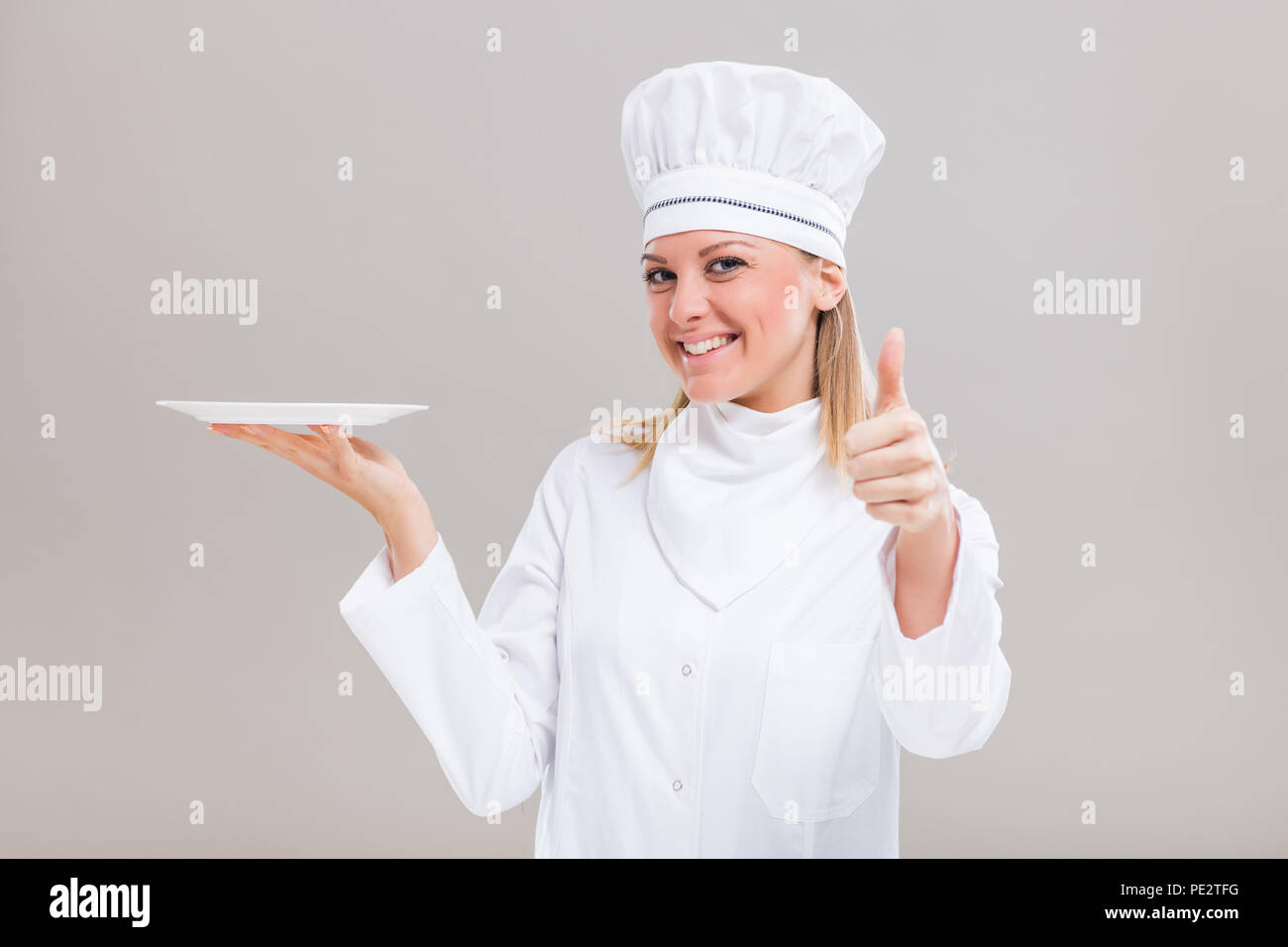 Bellissima femmina chef è la piastra di contenimento e mostrando il pollice su sfondo grigio. Foto Stock