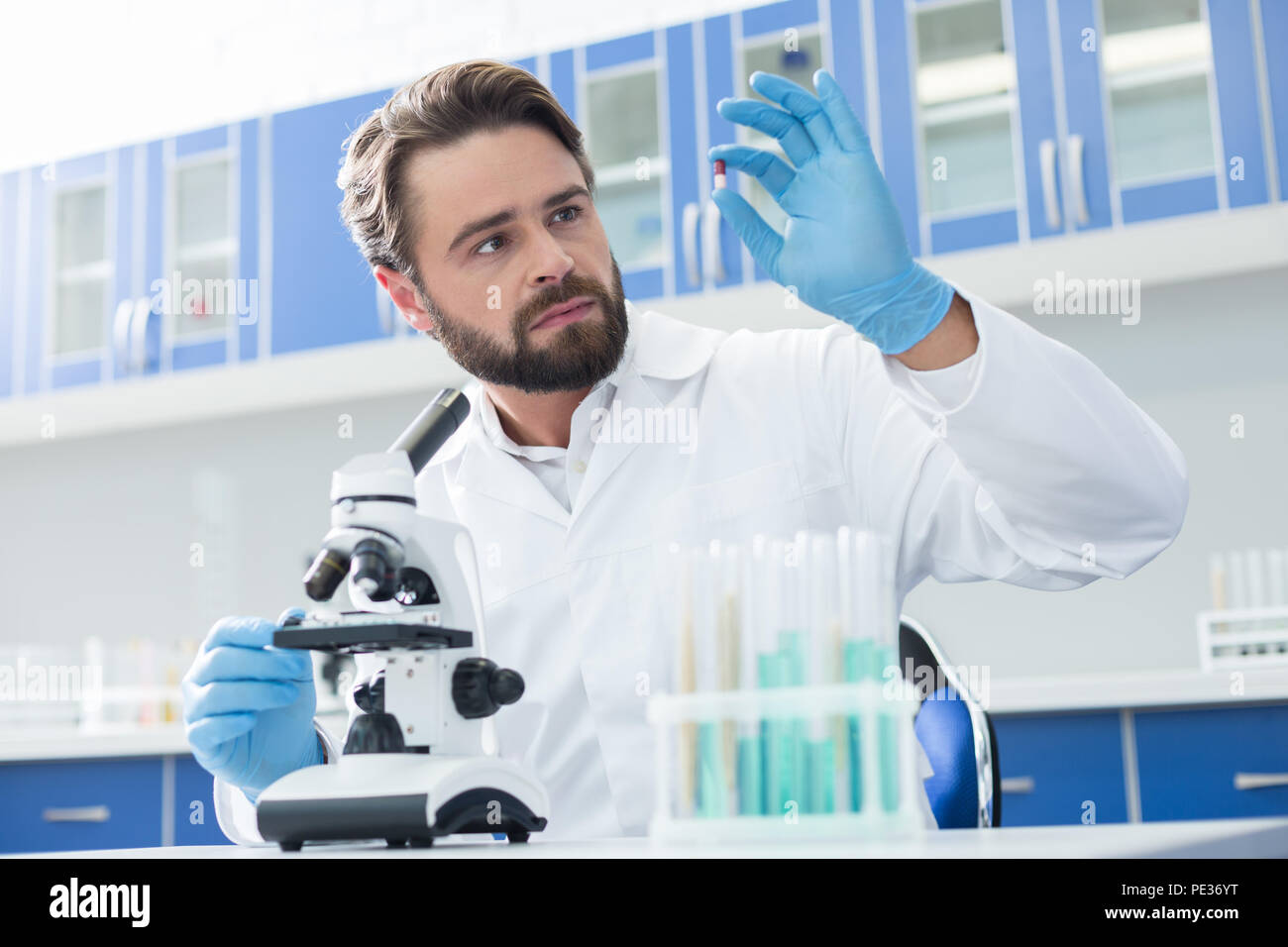 Nuove cure. Professional genius biologo barbuto tenendo una pillola e guardarlo mentre lo sviluppo di nuove cure Foto Stock