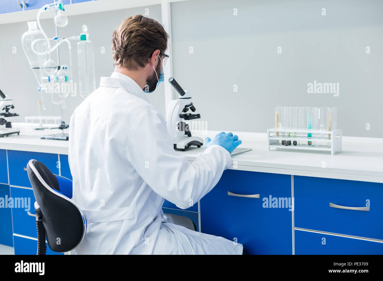 Professionale. Bello bello biologo maschile seduto sulla sedia e guardando nel microscopio durante il lavoro in laboratorio Foto Stock