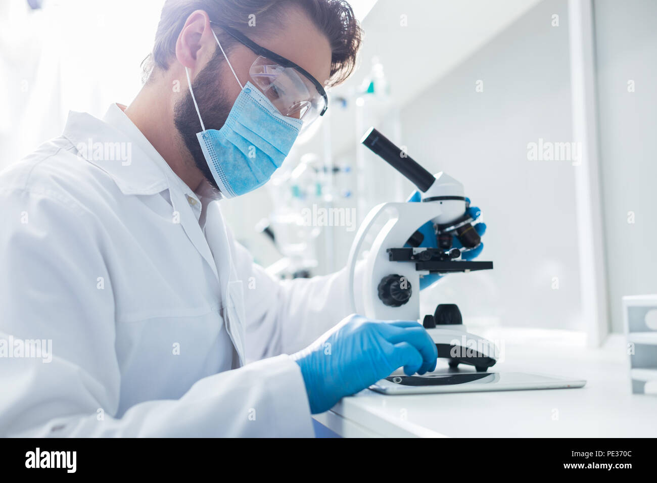 Laboratorio moderno. Smart bello bello scienziato seduti di fronte al microscopio e guardando il tablet schermo pur lavorando sulla sua ricerca Foto Stock