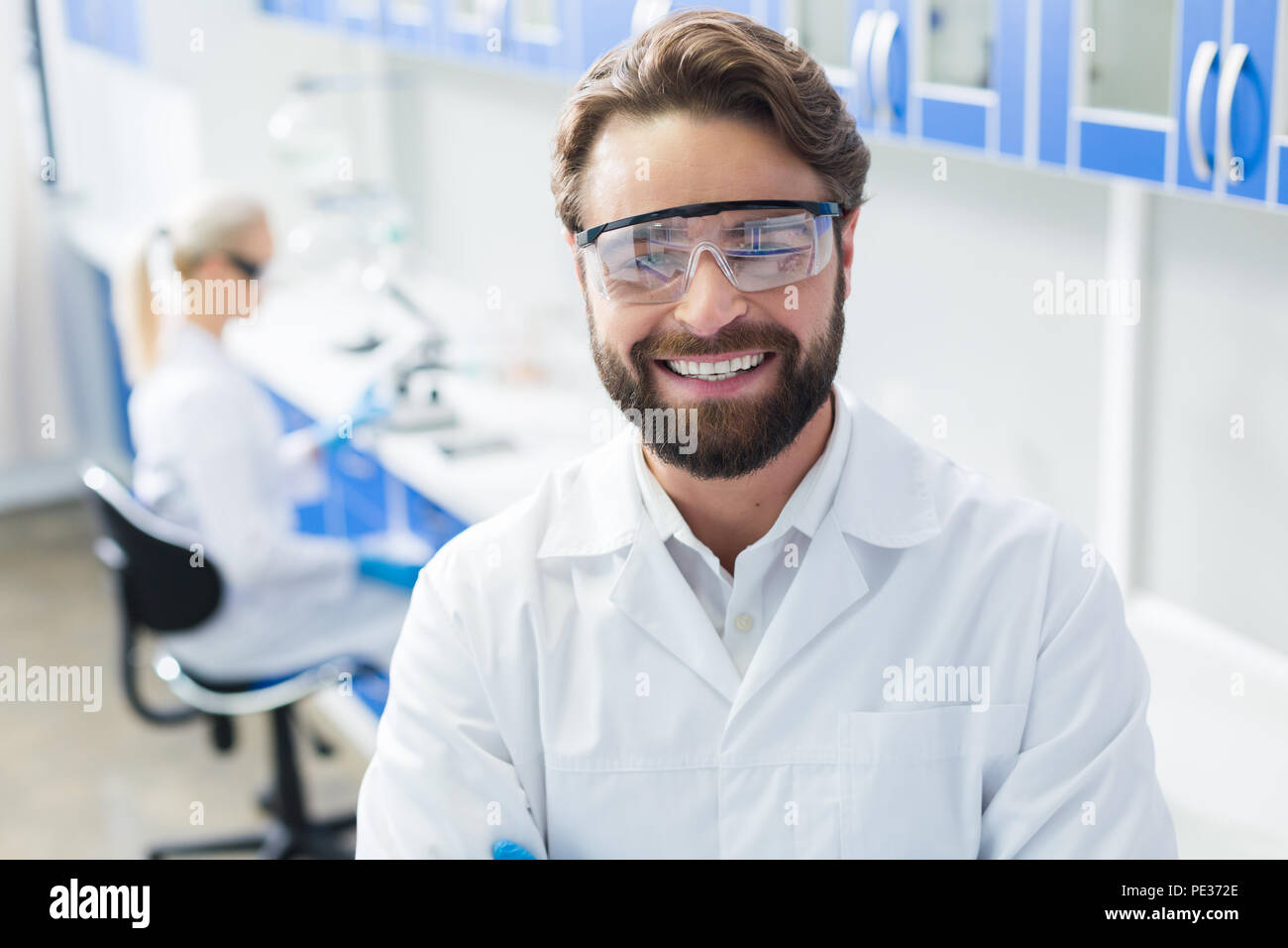 Occhiali di sicurezza. Felice positivo scienziato barbuto in piedi in laboratorio e indossare equipaggiamento protettivo durante il lavoro CI Foto Stock