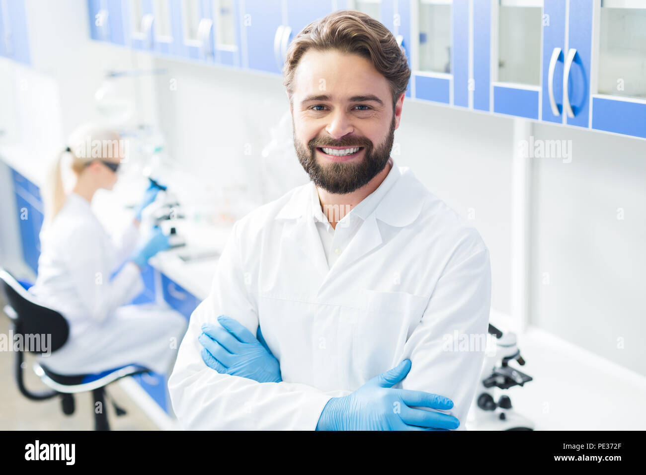 Laboratorio professionale. Smart fiducioso scienziato maschio croce permanente consegnato e sorridente durante il lavoro in laboratorio Foto Stock