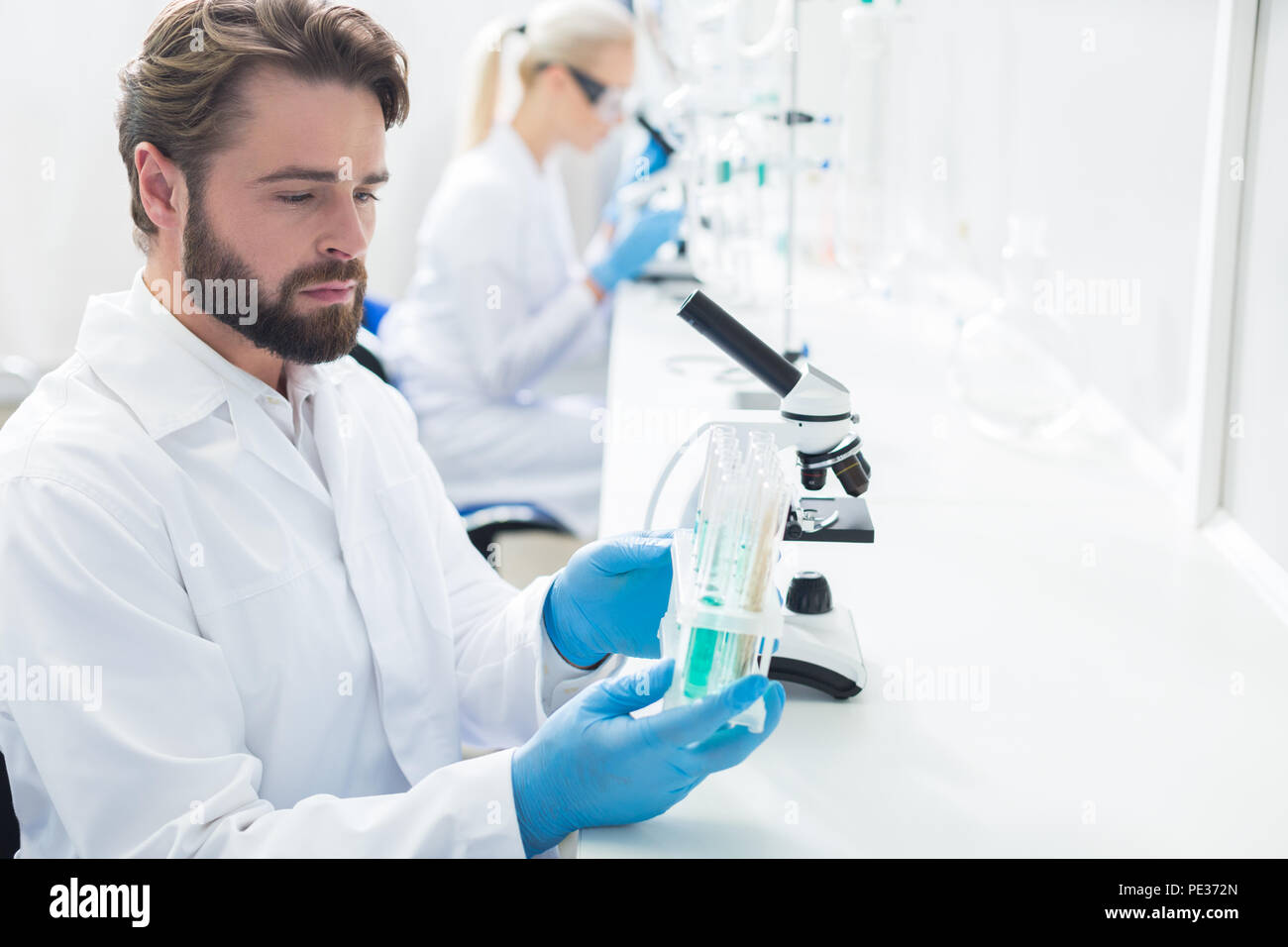 Modifiche scientifica. Smart piacevole di lavoro duro scienziato seduto in laboratorio e tenendo un rack con tubi di prova durante il corso di studi di loro Foto Stock
