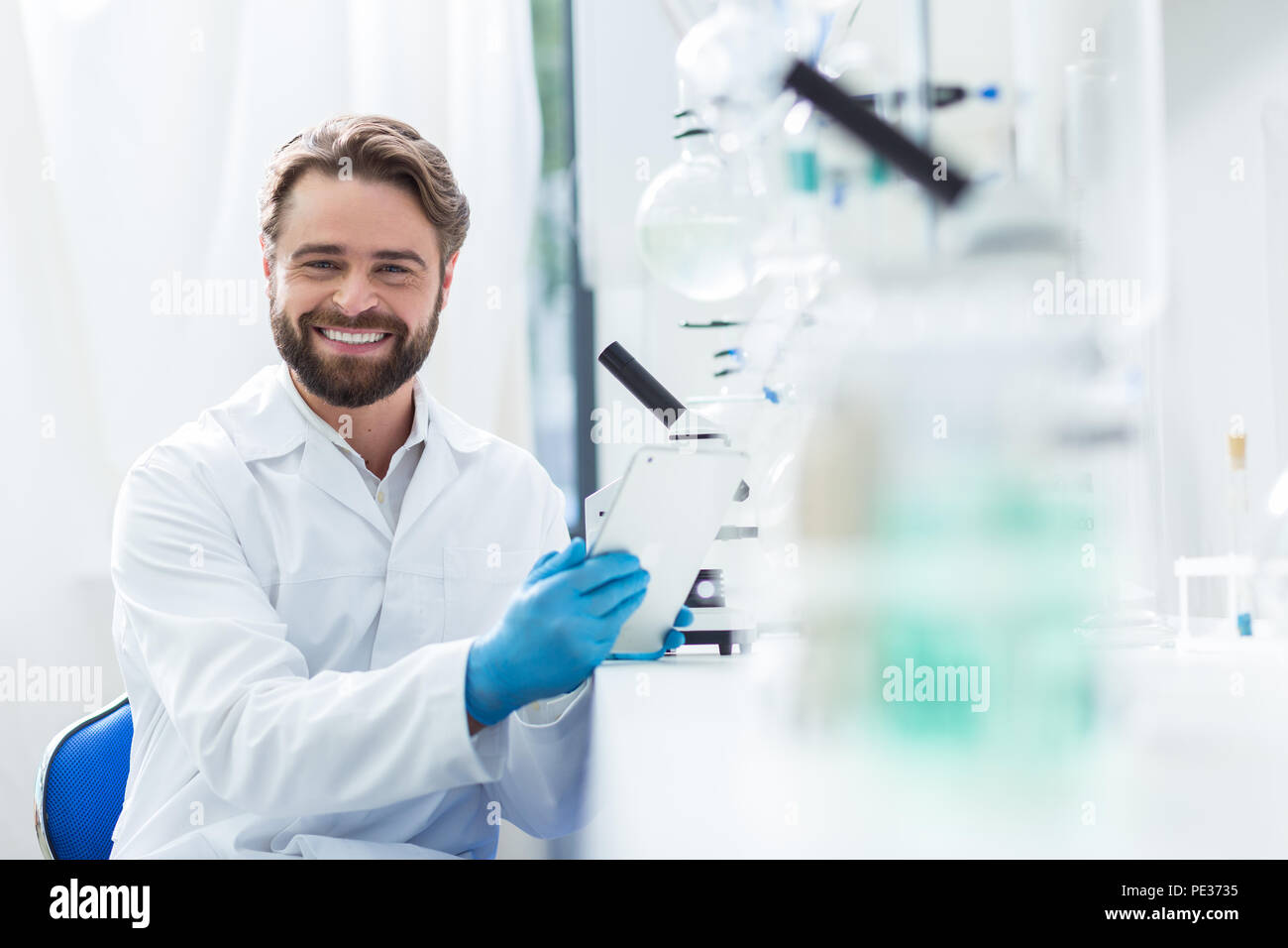 Stato d'animo positivo. Allegro nizza scienziato maschile seduto sulla sedia e sorridente a voi mentre sono felice circa il suo lavoro in laboratorio Foto Stock