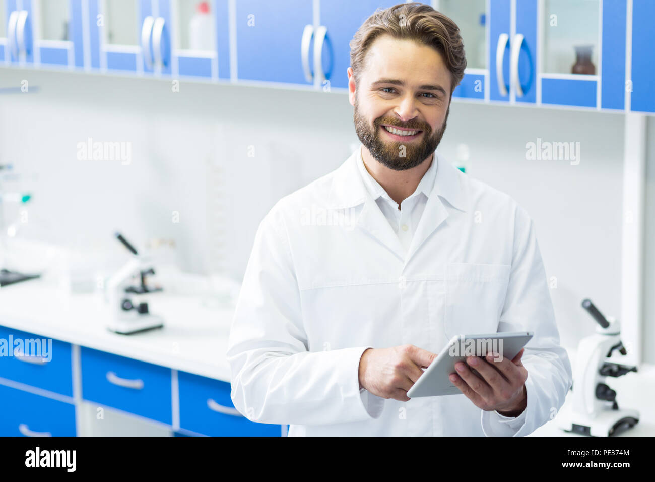 La tecnologia moderna. Smart positivo scienziato gioioso sorridente e utilizzando una compressa durante il lavoro nel laboratorio moderno Foto Stock