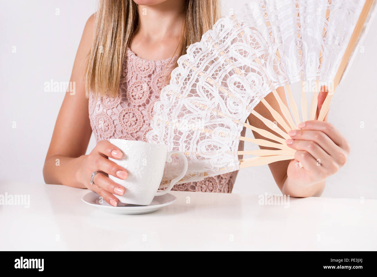 Bella giovane donna azienda tazza di caffè e pizzo bianco Ventilatore a mano sulla scrivania. Estate e concetto di calore. Close up, il fuoco selettivo Foto Stock