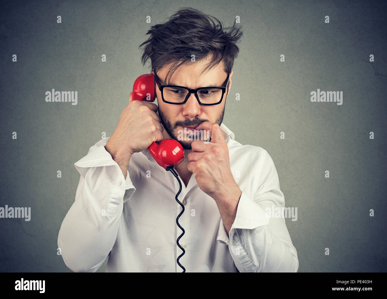Giovane uomo frustrato in bicchieri di chiamata sul telefono e mordere unghie nervoso cercando mentre si parla su sfondo grigio Foto Stock