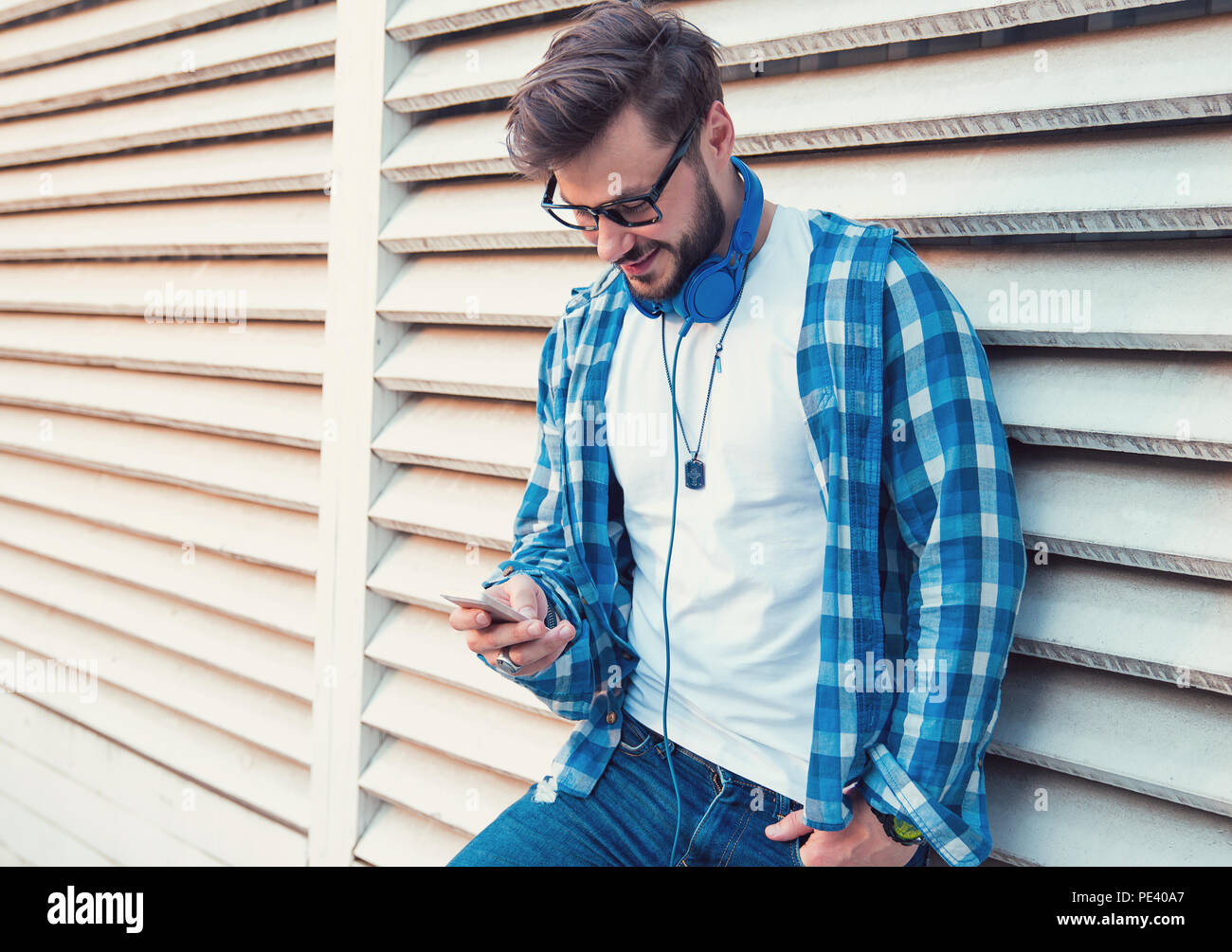 Tanga alla Moda Uomo in camicia blu e cuffie smartphone surf mentre appoggiata sulla parete su strada e sorridente Foto Stock