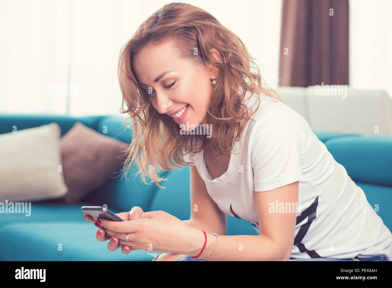 Felice ragazza casual scrivere messaggi su un telefono cellulare in appoggio su un lettino a casa nel suo accogliente appartamento Foto Stock
