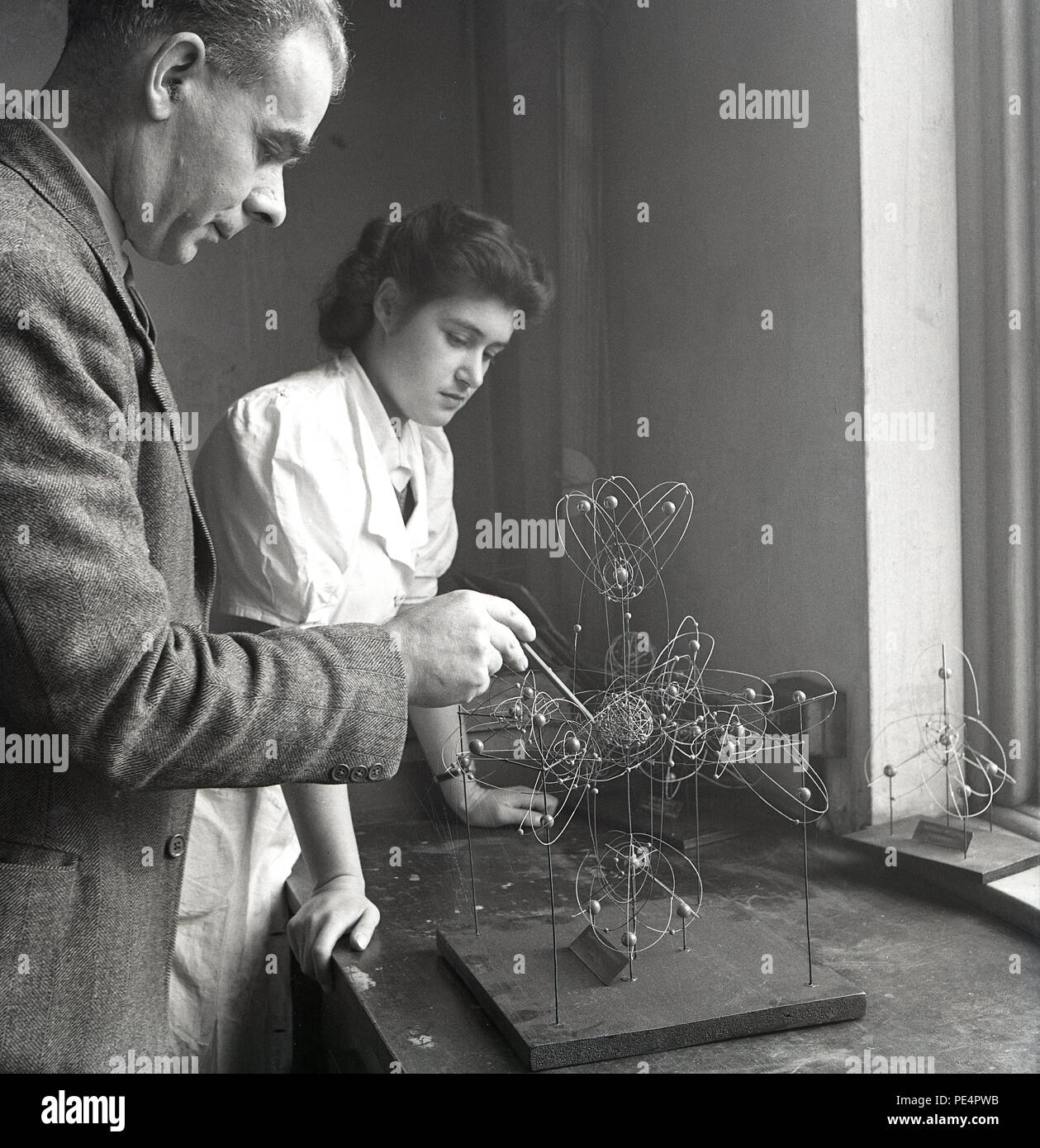Degli anni Cinquanta, maschio professore universitario di insegnare la scienza della fisica a un femlale studente utilizzando un penceil e un atomo di metallo del modello su una base di legno seduto su un banco di lavoro, Inghilterra, Regno Unito. Foto Stock