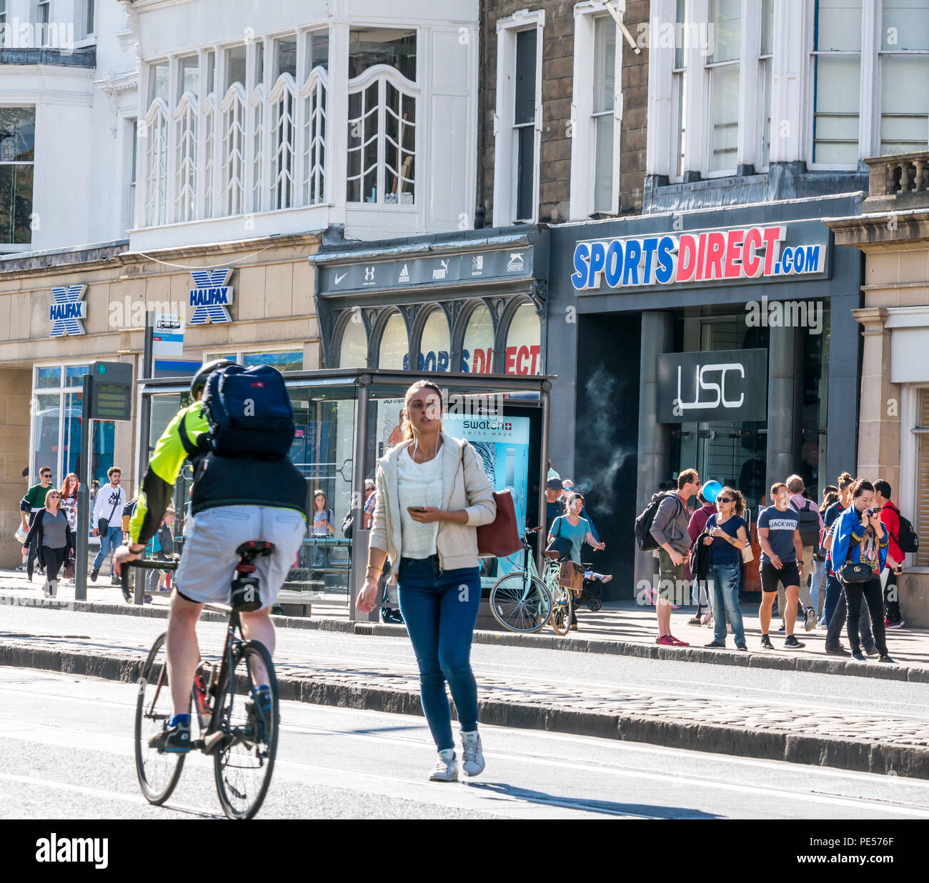 Donna di strada di attraversamento e il ciclista su Princes Street con sport diretto e USC shopfront e Halifax Building Society, Edimburgo, Scozia, Regno Unito Foto Stock