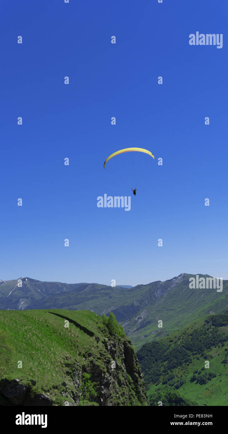 Un parapendio con persone vola sopra le cime della montagna in un luminoso cielo blu sullo sfondo di una gola di montagna. La vegetazione verde copre il mo Foto Stock