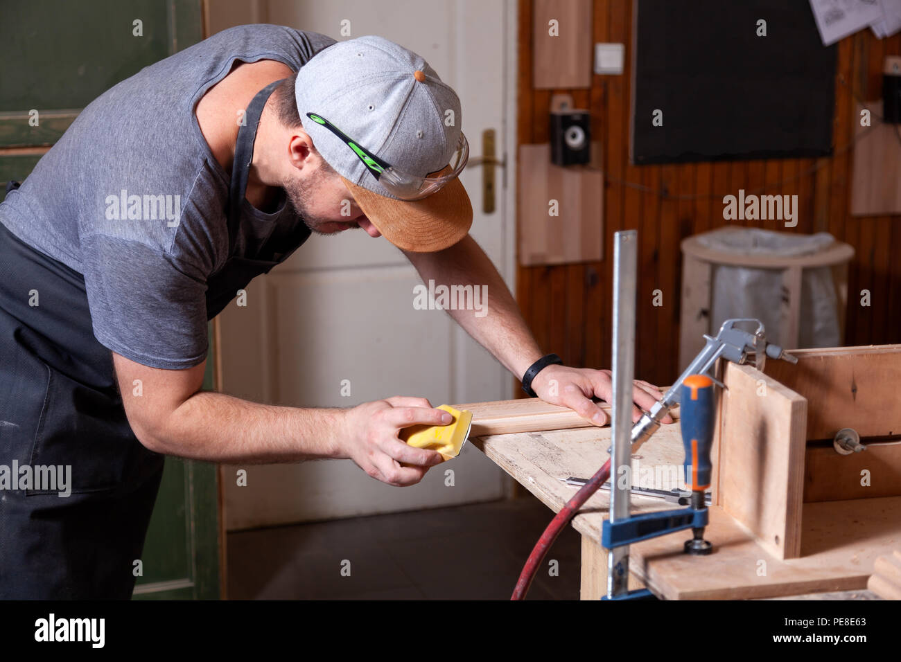 Close up del lavoro di un uomo in un cappuccio e camicia lucida il blocco di legno con carta vetrata prima della verniciatura in officina Foto Stock