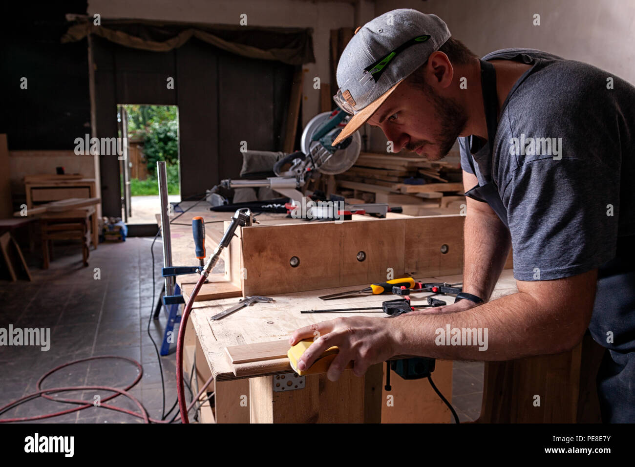 Close up del lavoro di un uomo in un cappuccio e camicia lucida il blocco di legno con carta vetrata prima della verniciatura in officina, in background, strumenti e d Foto Stock