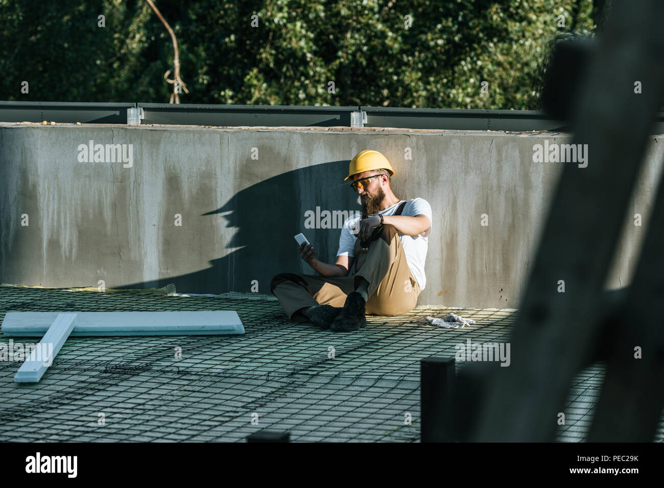 Giovani builder in hardhat e googles protezione utilizzando lo smartphone e in appoggio al sito in costruzione Foto Stock