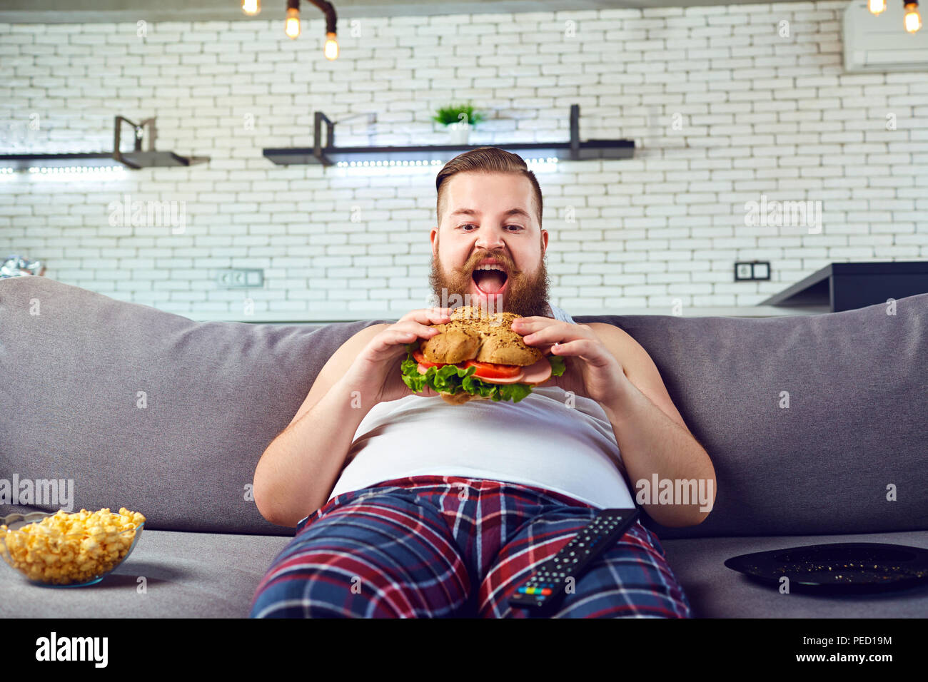 Fat funny man in pigiama mangiare un hamburger sul divano di casa. Foto Stock