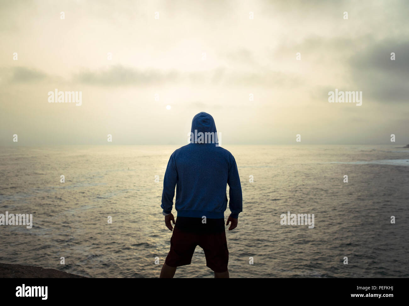 Silhouette di un uomo in una felpa con cappuccio da dietro a guardare il tramonto dorato oltre oceano. Punta Hermosa surfisti (città), Provincia di Lima, Perù. Lug 2018 Foto Stock