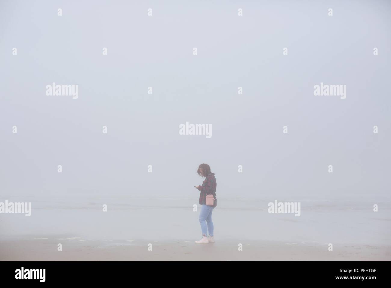 Una giovane donna che guarda il telefono mentre è in piedi su una spiaggia di nebbia vicino Firenze, Oregon, Stati Uniti d'America. Foto Stock