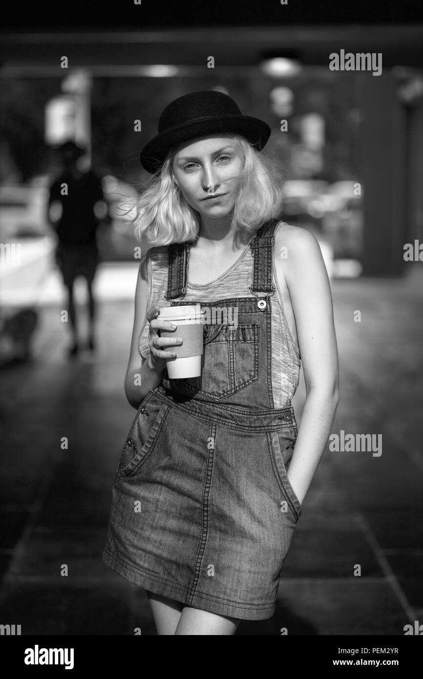 Ritratto in bianco e nero del Caucaso giovane adolescente bionda modello alternativo Ragazza donna tshirt, jeans romper cercando nella fotocamera tenendo tazza di caffè Foto Stock