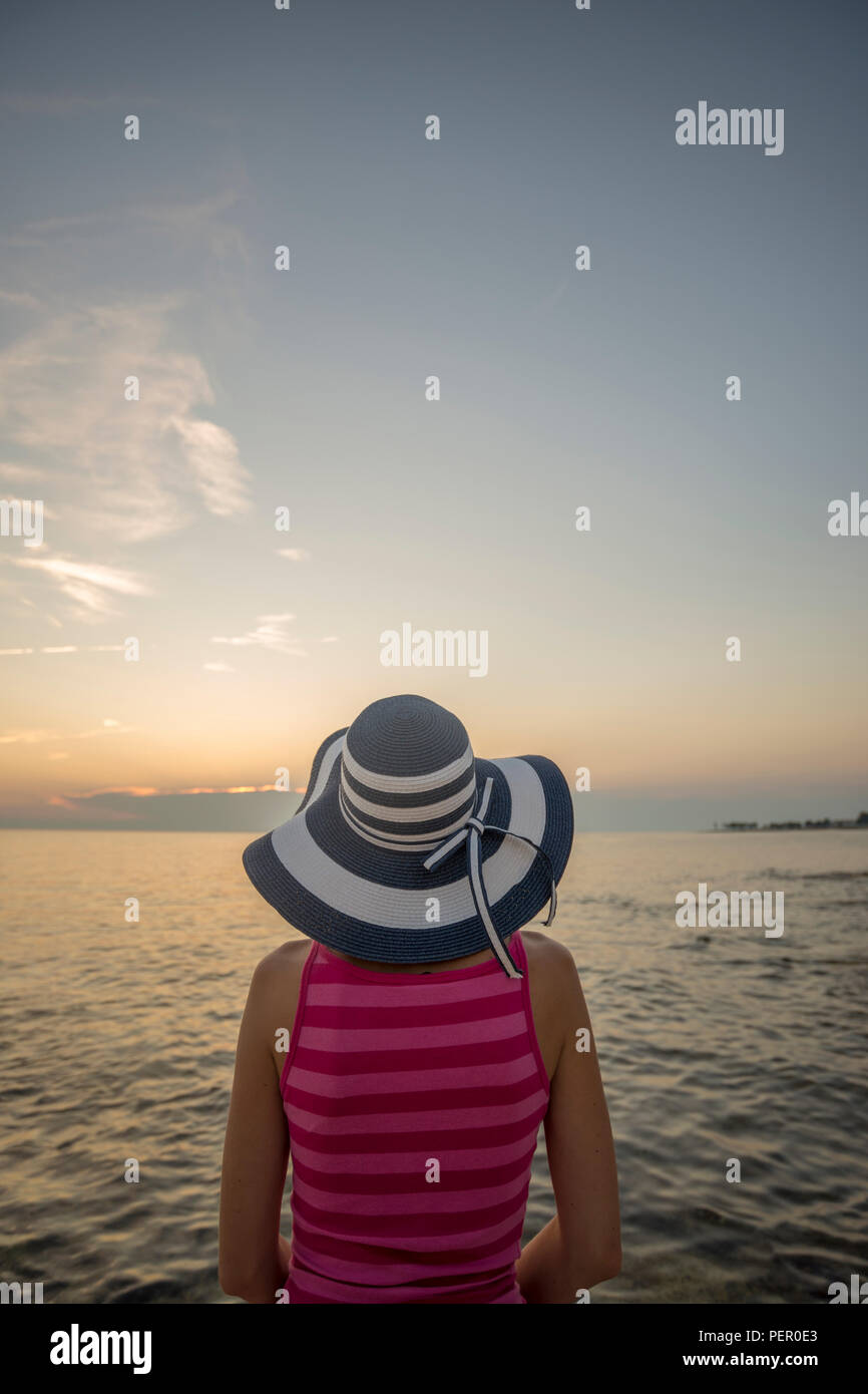 Vista posteriore della donna in rosa camicia senza maniche e a larga tesa a guardare il tramonto sul mare, copia dello spazio. Foto Stock