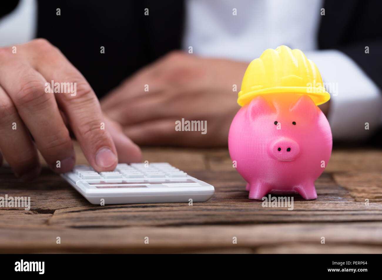 Mano umana utilizzando la calcolatrice accanto a piggybank con giallo hardhat sulla scrivania in legno Foto Stock