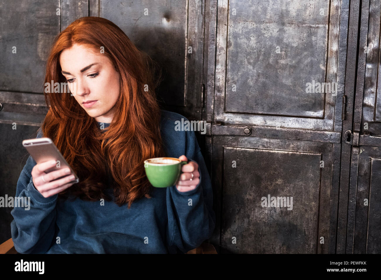 Giovane donna con lunghi capelli rossi seduta a tavola, tenendo tazza di caffè, controllando il suo telefono cellulare. Foto Stock