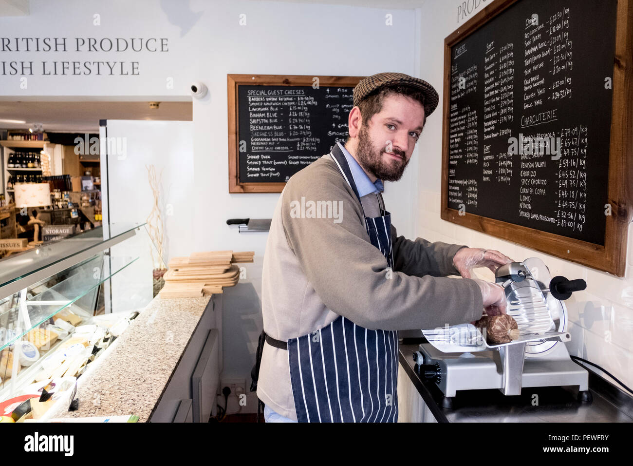 Uomo Barbuto che indossa il tappo piatto e grembiule permanente al contatore in un negozio di specialità gastronomiche, guardando la fotocamera. Foto Stock