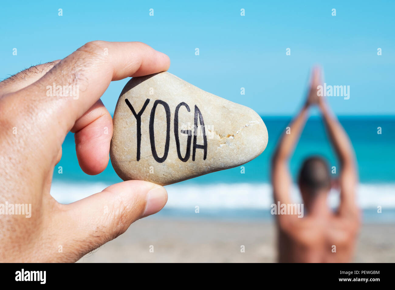 Primo piano della mano di un giovane uomo caucasico sulla spiaggia tenendo una pietra con lo yoga scritto in esso, e un giovane uomo caucasico la pratica dello yoga in th Foto Stock
