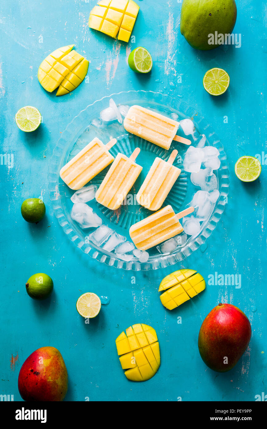 Mango popsicles su una lastra di vetro con cubetti di ghiaccio, lime e mango fruit,vista superiore, scheda blu. Foto Stock