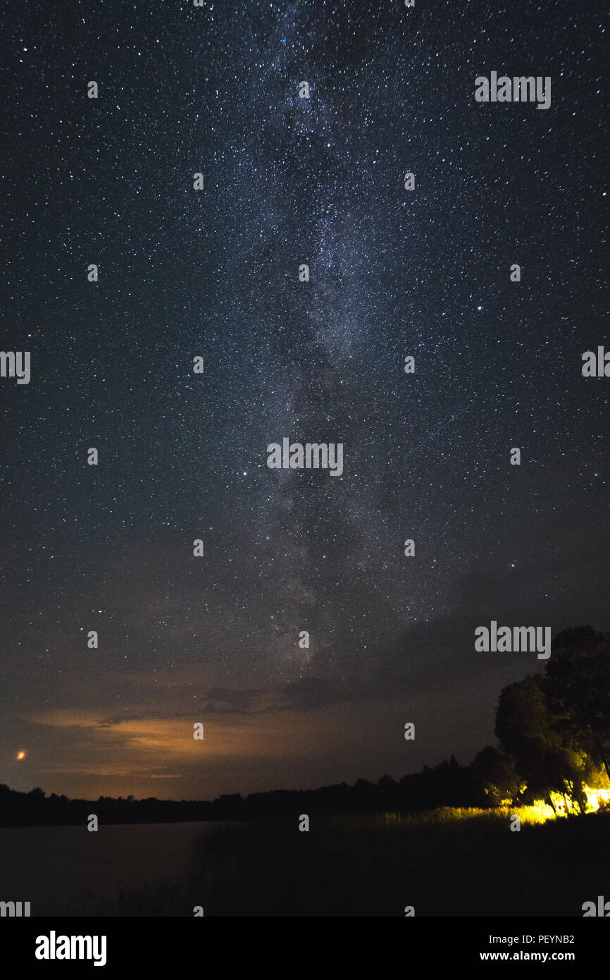 Via Lattea e cielo stellato notturno sul lago calmo con sagome forestali, raggio laser verde e riflessi celesti in un paesaggio naturale buio Foto Stock