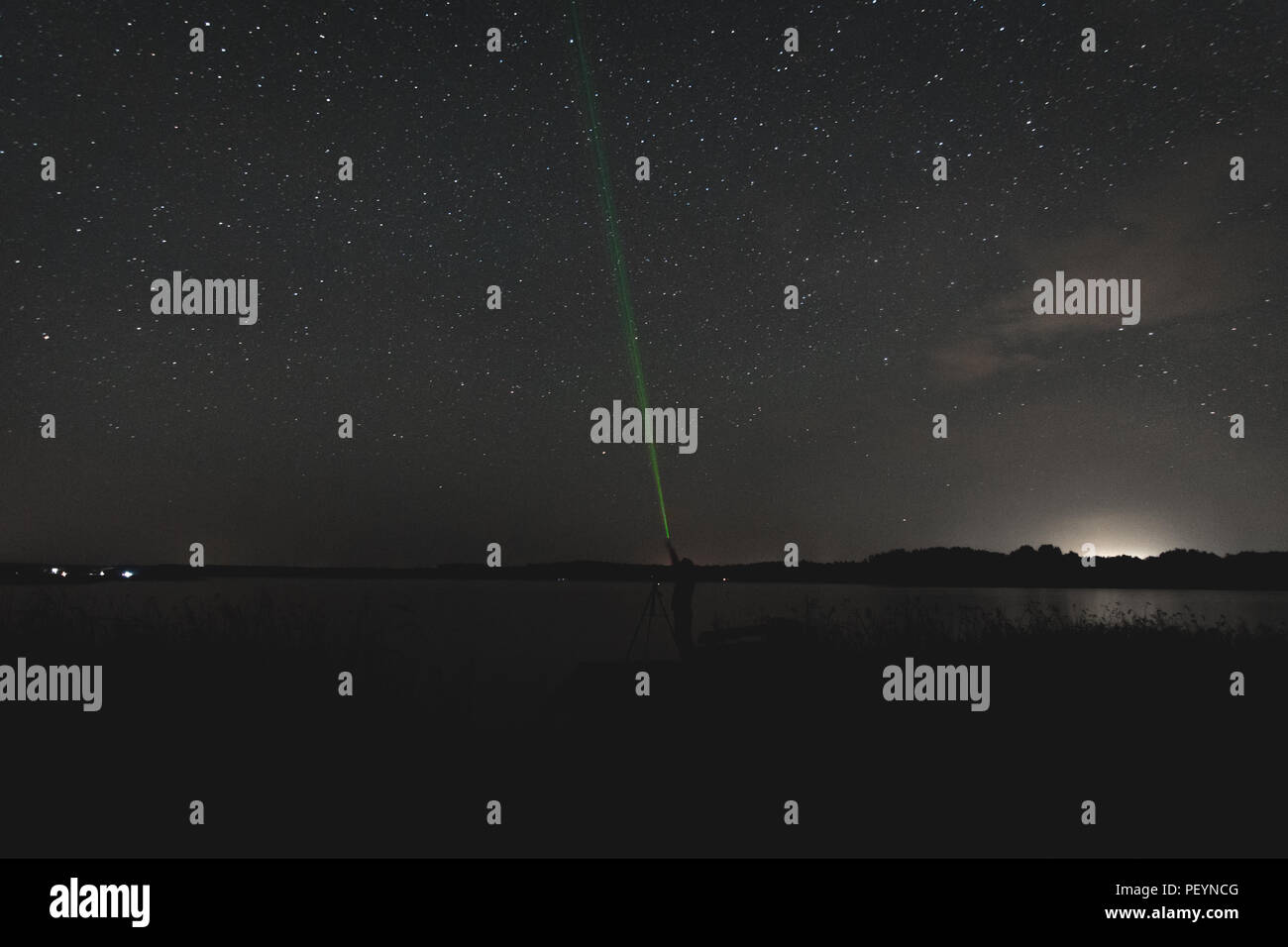 Via Lattea e cielo stellato notturno sul lago calmo con sagome forestali, raggio laser verde e riflessi celesti in un paesaggio naturale buio Foto Stock