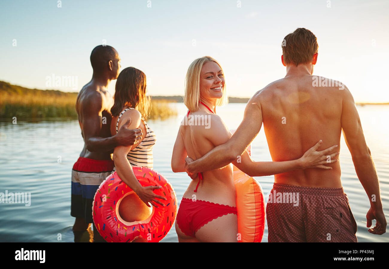 Donna sorridente in un bikini guardando sopra la sua spalla mentre in piedi con il suo fidanzato e un altro giovane in un lago di guardare il tramonto Foto Stock