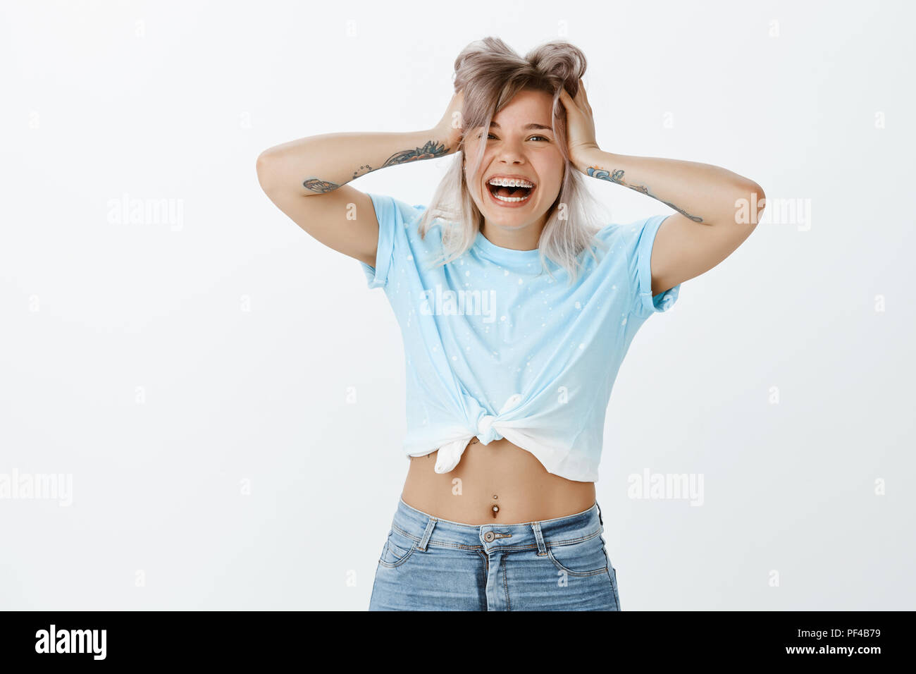 Piscina colpo di emtoive gioiosa unione femmina con bracci tatuati e forato ventre, sorridente largamente e tenendo le mani sui capelli, essendo sorpreso ed entusiasmato da grande incredibile news oltre il muro grigio Foto Stock