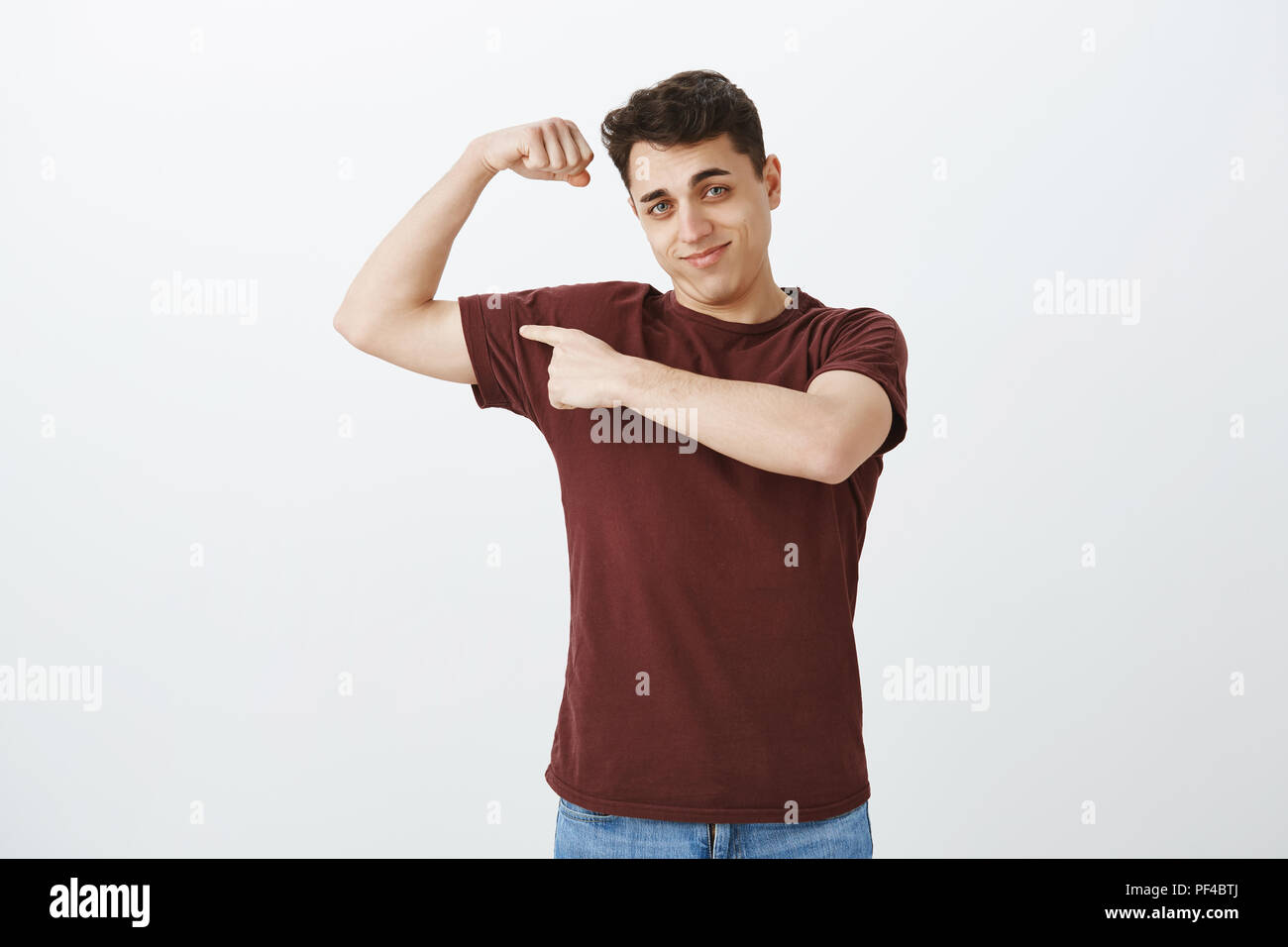 Ritratto di bello felice maschile ragazzo in t-shirt rossa, alzando la mano e mostra i muscoli, puntando al braccio con il dito indice, vantarsi circa la resistenza del corpo dopo la visita di palestra, godendo di lavoro Foto Stock