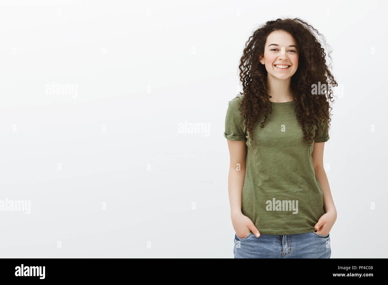 Studio shot del timido carino curly-pelose studentessa in verde scuro e t-shirt, tenendo le mani nelle tasche e sorridente largamente mentre la sensazione di disagio e gioiosi nella nuova società oltre il muro grigio Foto Stock